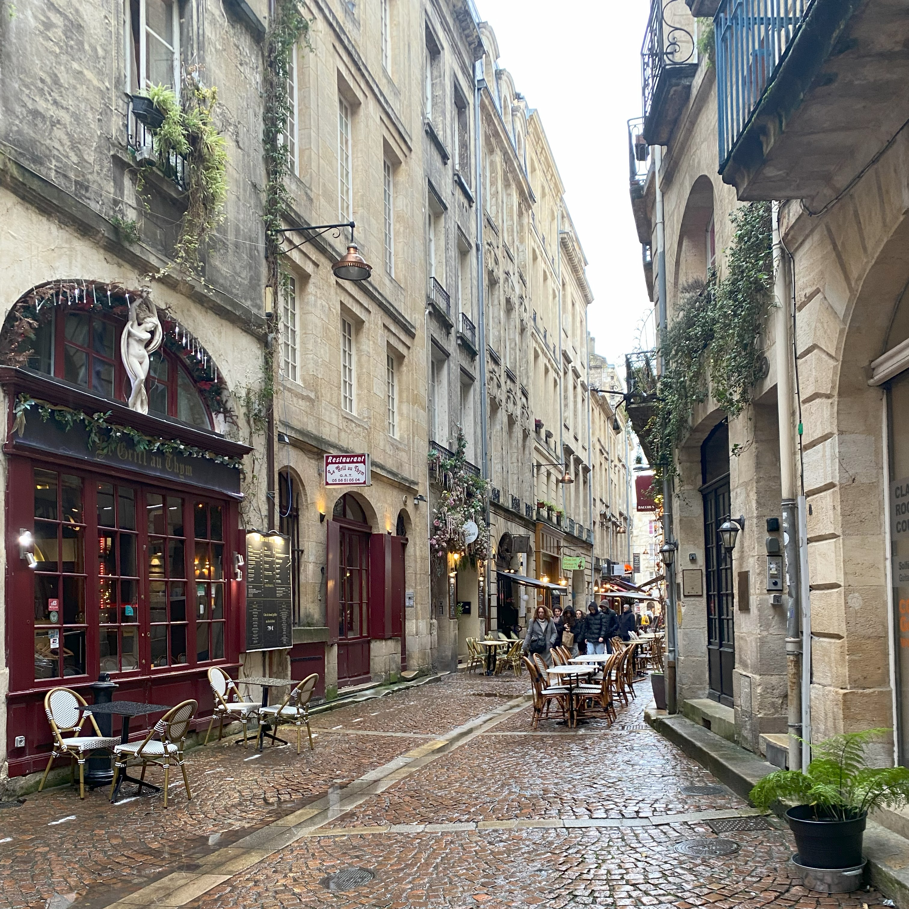 Rua estreita e histórica com paralelepípedos, prédios de pedra e mesas de café nas calçadas.